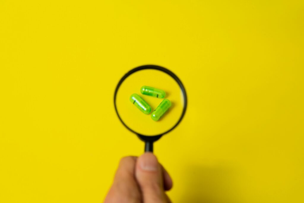 Top view of crop unrecognizable person holding magnifier over green pills placed on bright yellow surface in studio